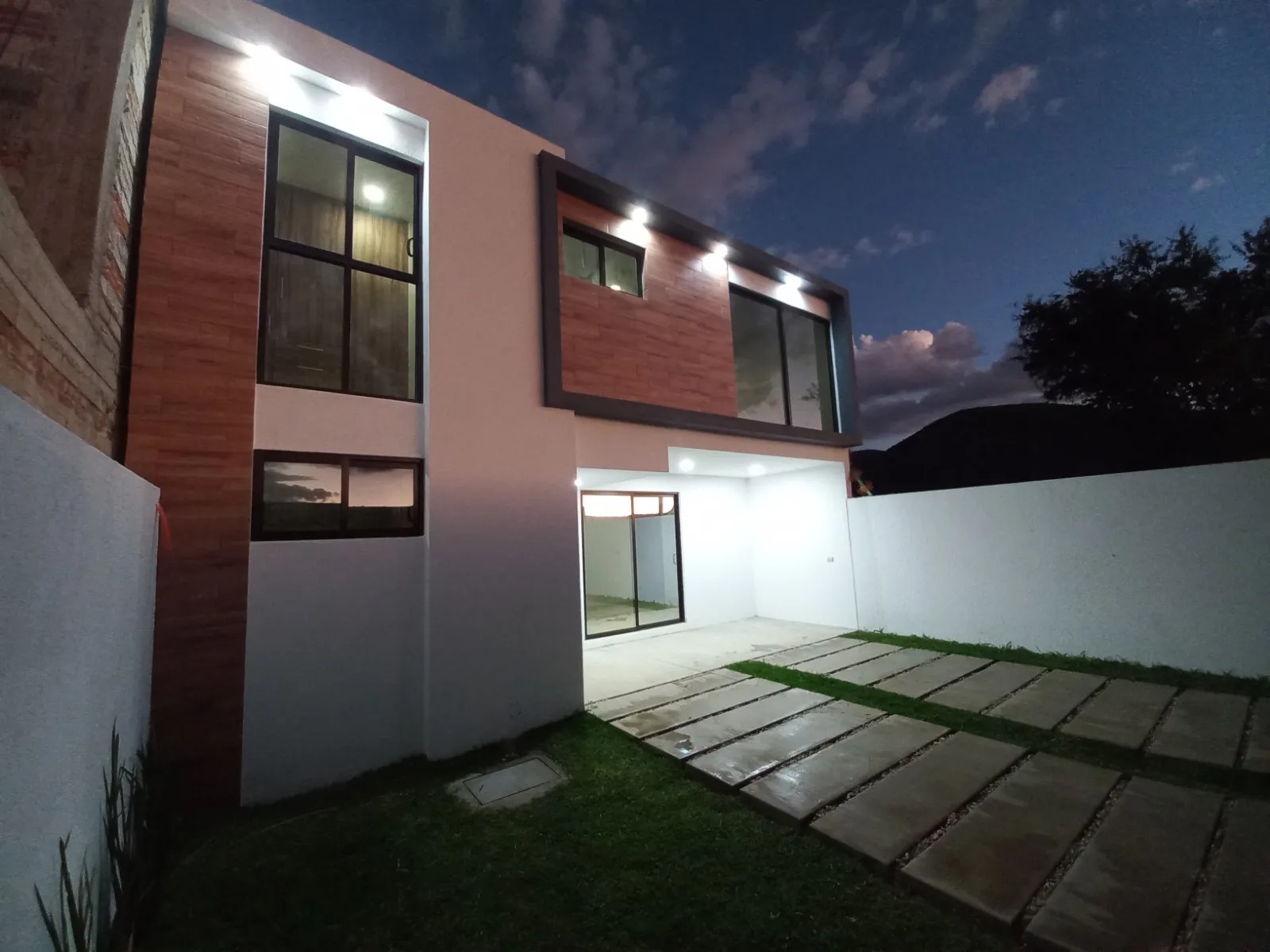 Casa de dos plantas al anochecer, con paneles de madera y patio con andador